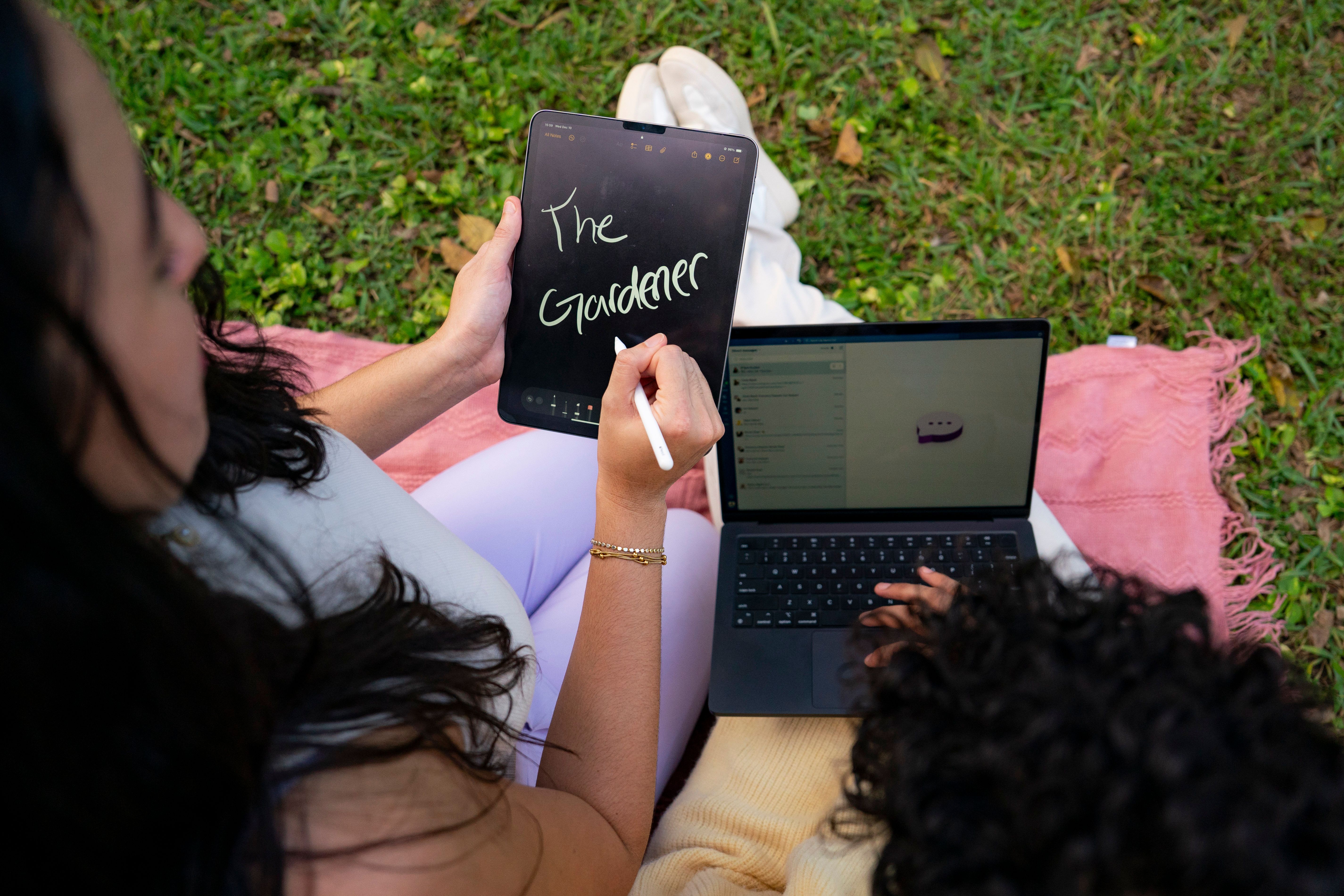 A creative team member writing “The Gardener” on a tablet with a stylus while another team member works on a laptop outdoors.